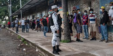 Migrantes haciendo fila vigilados por una agente de la Guardia Nacional